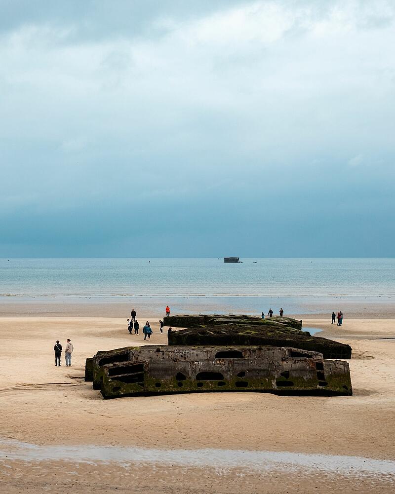 [Translate to Anglais:] Wide shot of a sandy beach under an overcast sky, where tourists walk around large concrete blocks, an excursion easily accessible from Les Castels campings in Normandy.