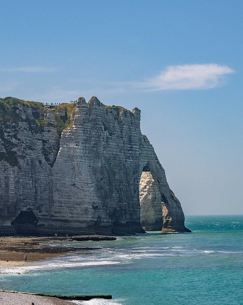 [Translate to Anglais:] A vertical shot of the famous towering white chalk cliffs and natural arch of Étretat, with waves crashing onto the beach, located near a Les Castels camping resort.