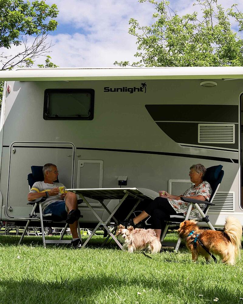 [Translate to Anglais:] A man and a woman sitting at a camping table under the awning of a modern Sunlight motorhome on a pet-friendly pitch at Les Castels.