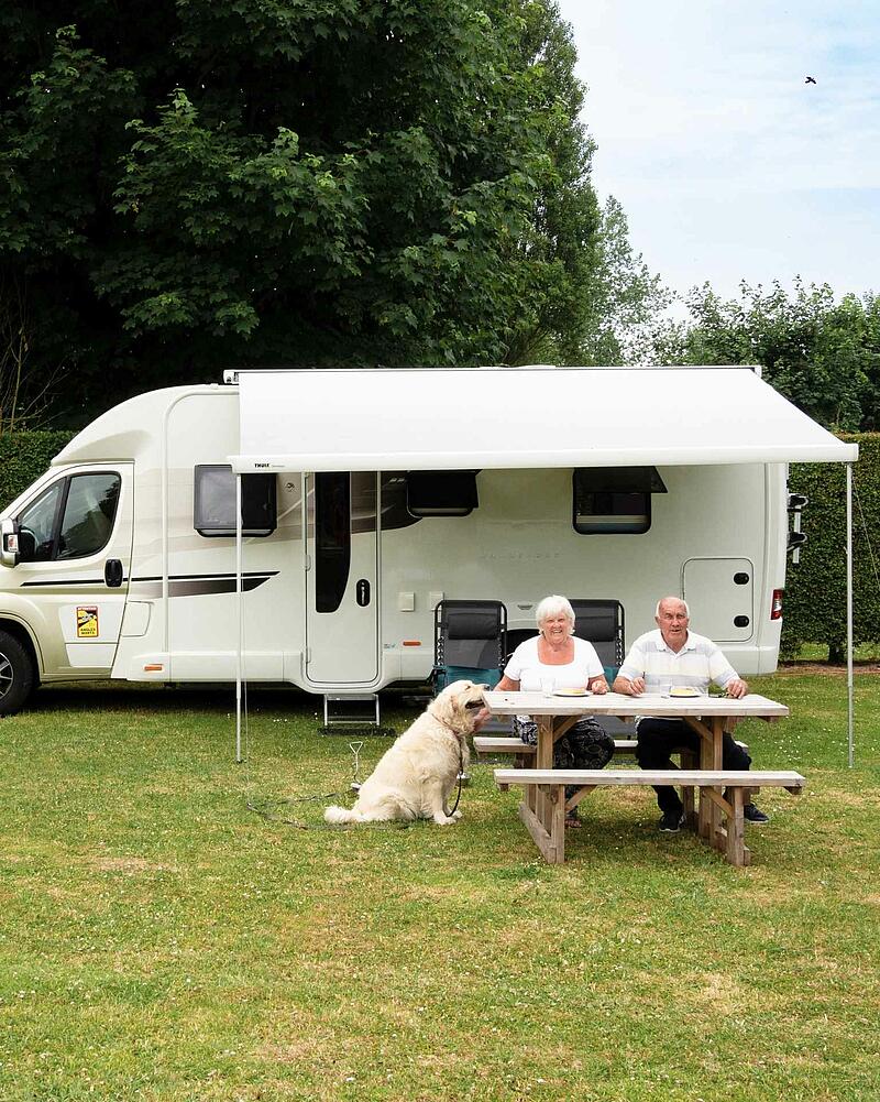 [Translate to Anglais:] A senior couple enjoying a meal at a wooden picnic table outside their white motorhome under an awning, accompanied by their golden retriever dog on a green campsite pitch.