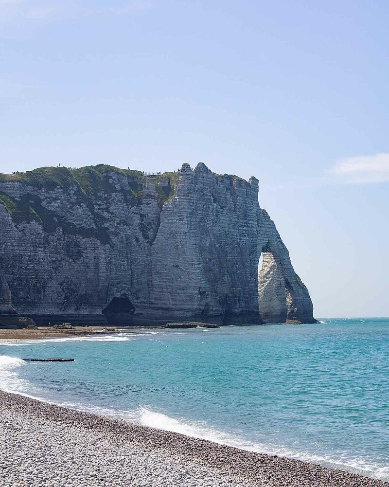 [Translate to Anglais:] The dramatic white chalk cliffs and natural rock arch of Étretat rising above a pebble beach and the turquoise waters of the sea in Normandy.
