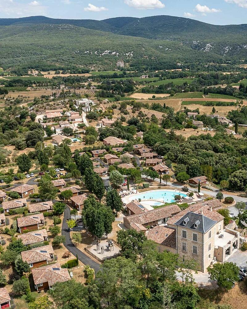 [Translate to Anglais:] Drone perspective of the heated outdoor swimming pool, stone estate building, and eco-friendly wooden chalets surrounded by nature at a 5-star Les Castels campsite.