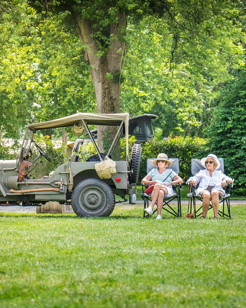 [Translate to Anglais:] Two senior women wearing sun hats sitting on camping chairs on a green lawn next to a classic green vintage military jeep at a Les Castels campsite.