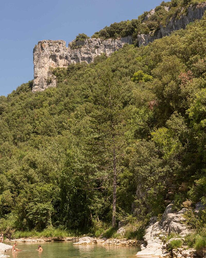 [Translate to Anglais:] People swimming and relaxing in the clear river water at the foot of towering limestone cliffs in the Ardeche Gorges near a luxury Les Castels estate.