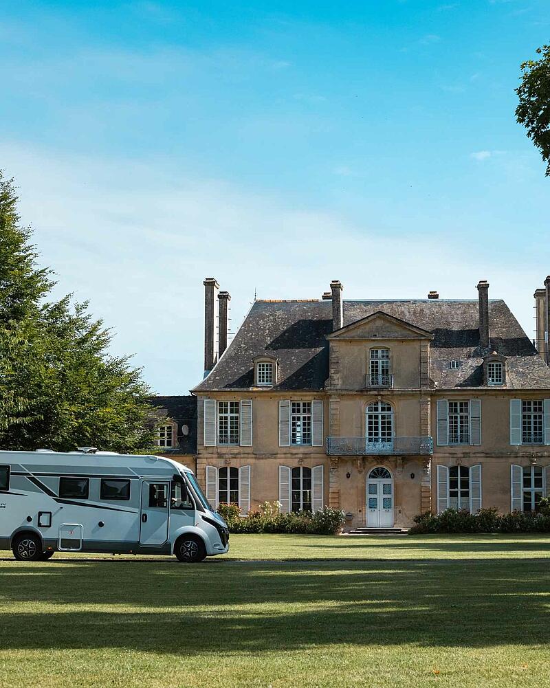 [Translate to Anglais:] A sleek white motorhome parked on the manicured lawns in front of a grand, historic French manor house during a summer road trip.