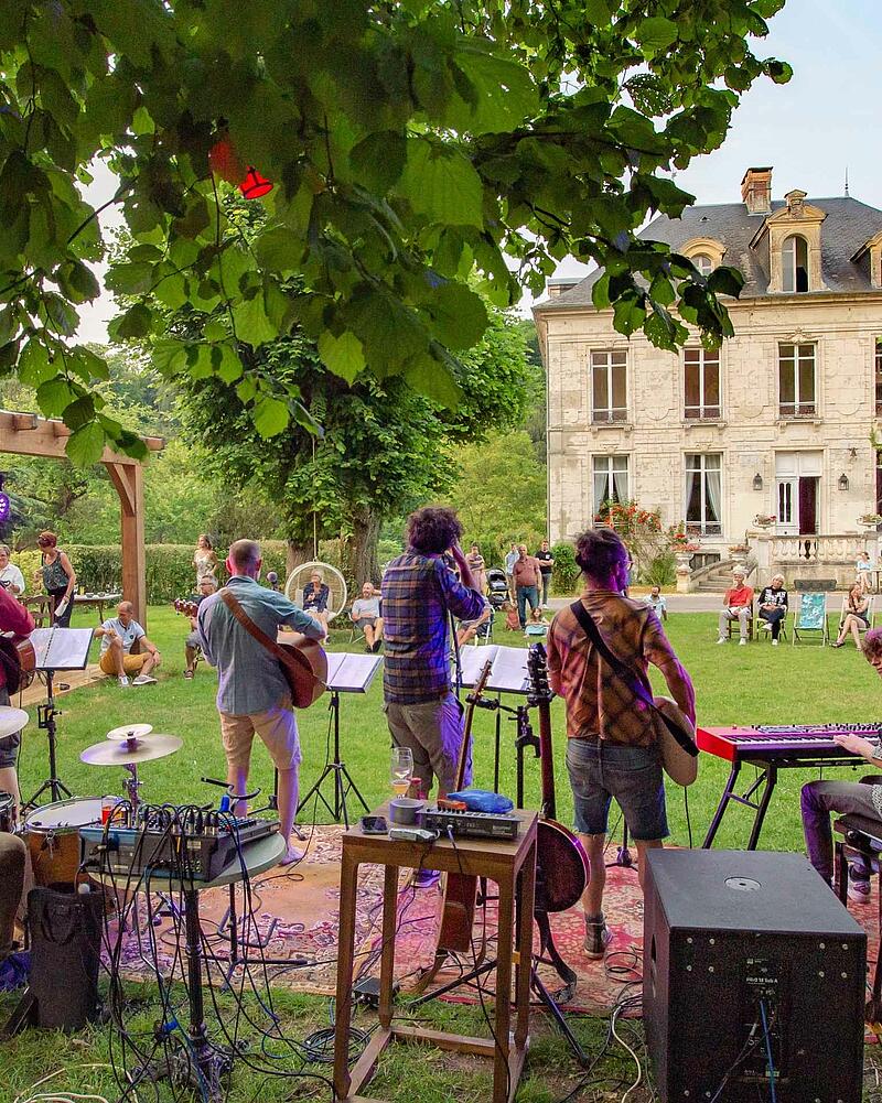 [Translate to Anglais:] Stage view from behind a live band playing guitars and keyboards outdoors for an audience seated on the grass during a premium evening event at Les Castels.