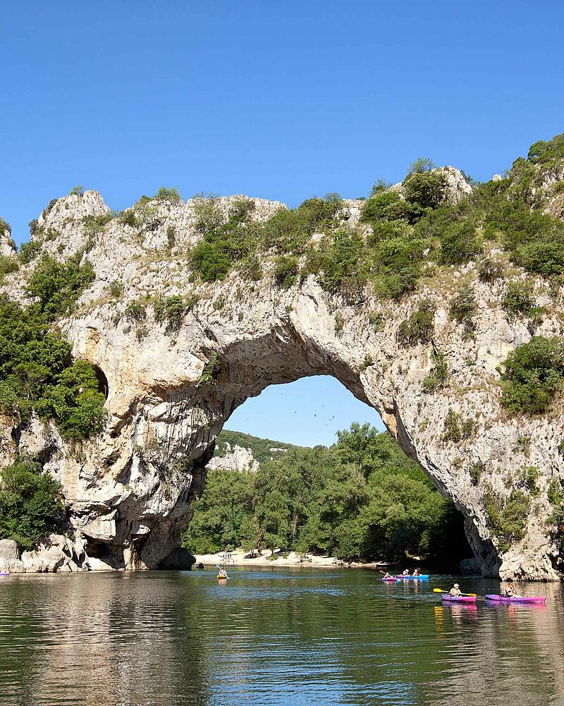 [Translate to Anglais:] Colorful holidaymakers paddling on kayaks and canoes on the river right underneath the giant natural stone bridge of Pont d Arc near Les Castels luxury campsites.