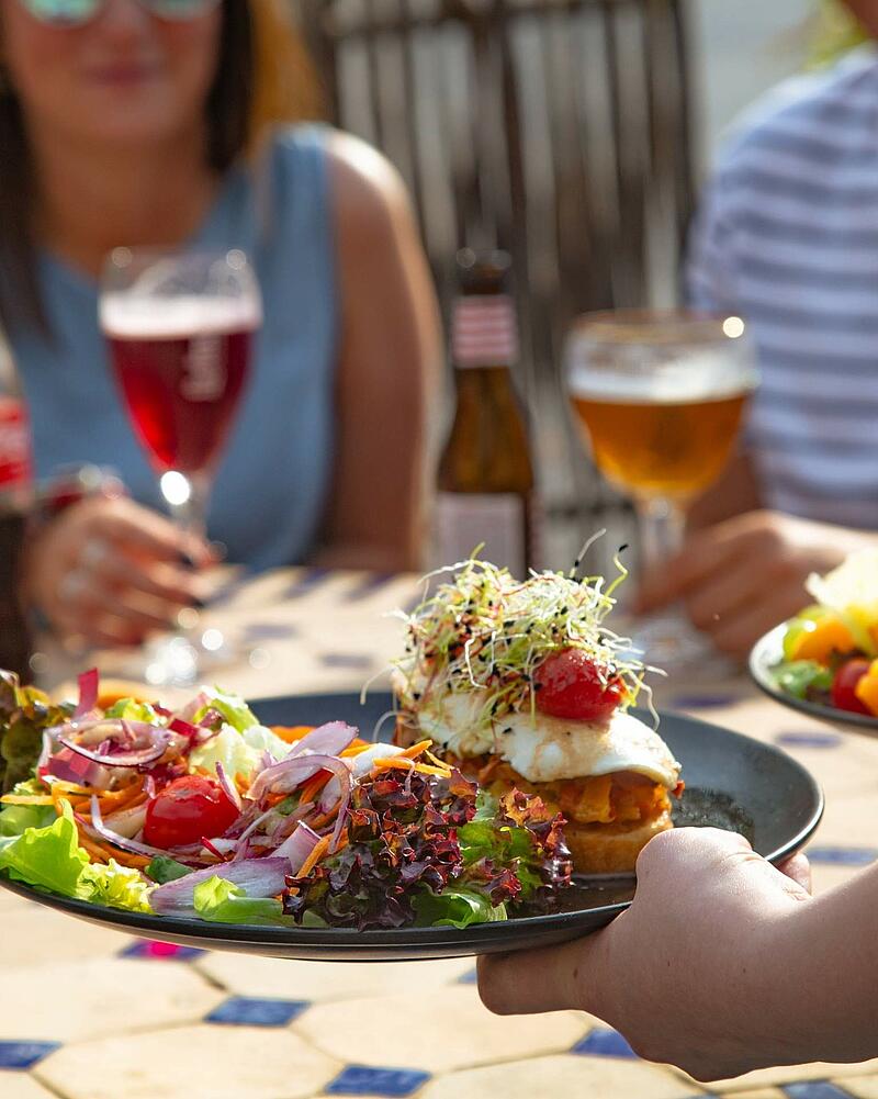 [Translate to Anglais:] A waiter serving a fresh, colorful salad platter with sprouts and a fried egg on toast at an outdoor terrace, with campsite guests enjoying beer and wine in the blurred background.