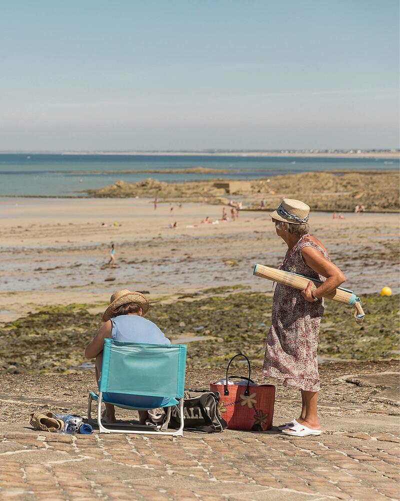 [Translate to Anglais:] Two women wearing straw hats spending a sunny day on a stone seawall, overlooking a vast sandy beach during a seaside holiday with Les Castels campings.