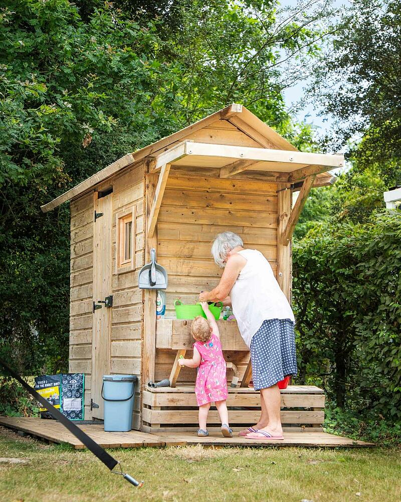 [Translate to Anglais:] Woman and young girl interact at a rustic wooden outdoor kitchen station on a Les Castels camping estate.