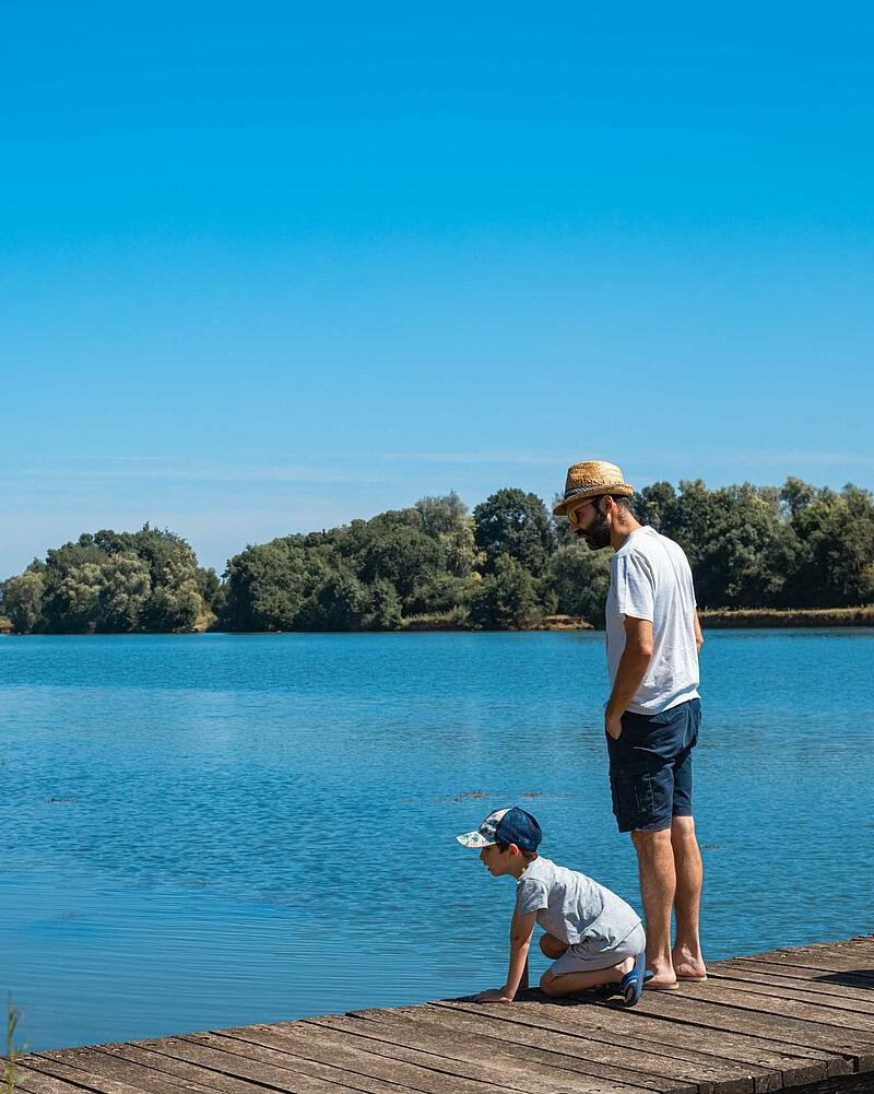 [Translate to Anglais:] A father wearing a straw hat stands next to his young son who is looking into the calm blue water from a wooden jetty on a private campsite lake.