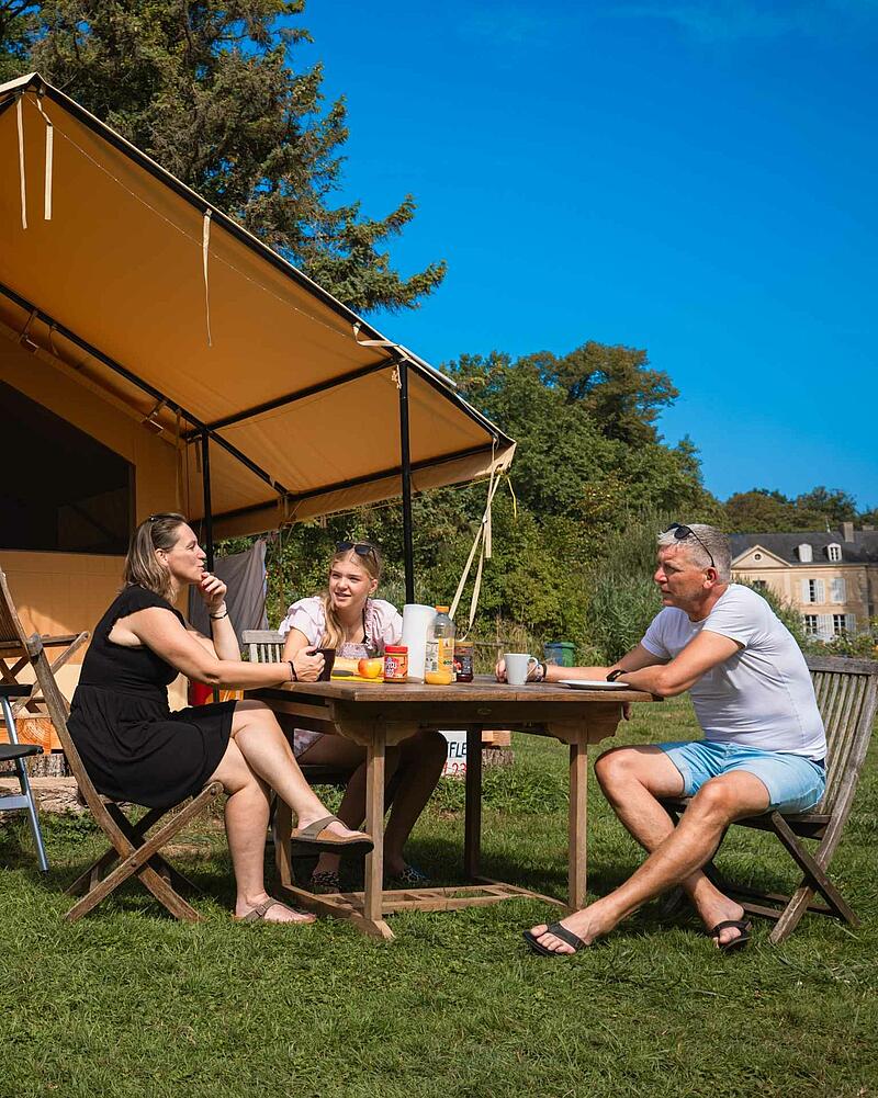 [Translate to Anglais:] A mother, father, and daughter sit around a wooden picnic table having breakfast outside a large luxury canvas glamping safari tent on a sunny campsite lawn.