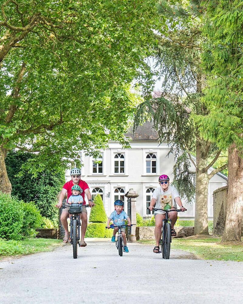 [Translate to Anglais:] A family of four, including two young children wearing helmets, cycling towards the camera on a gravel path during their holiday at a Les Castels domain.