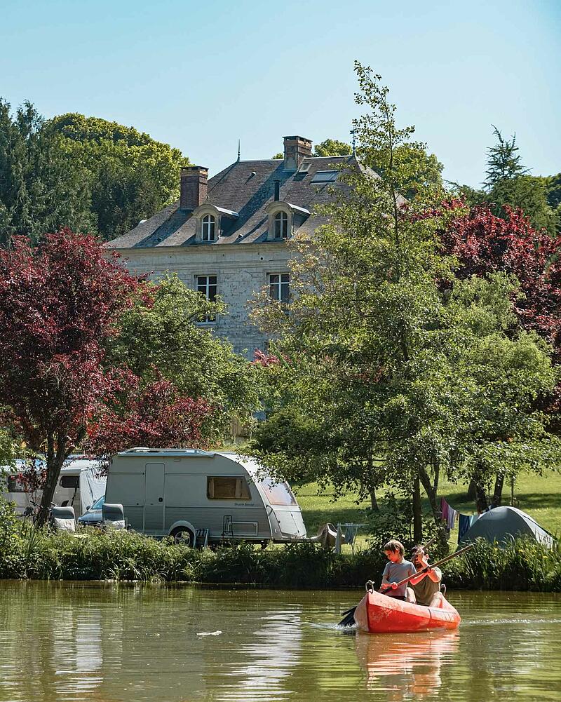 [Translate to Anglais:] An orange canoe with a man and a young boy paddling on a calm lake, with a caravan parked on the grassy bank of a Les Castels estate.