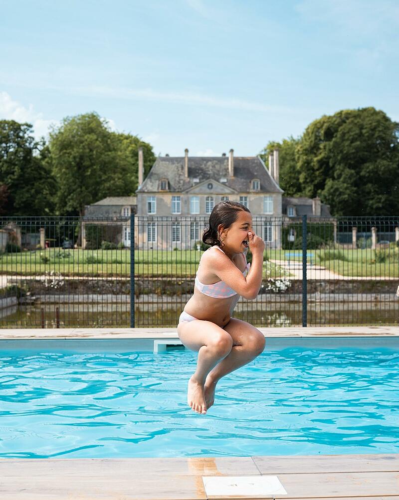 [Translate to Anglais:] A young girl mid-air in a cannonball jump into a crystal-clear outdoor swimming pool, located in the gardens of a grand French estate at Les Castels.