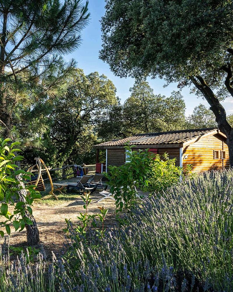 A cozy wooden chalet nestled among green trees and blooming purple lavender bushes at Castels camping Domaine de Sévenier.