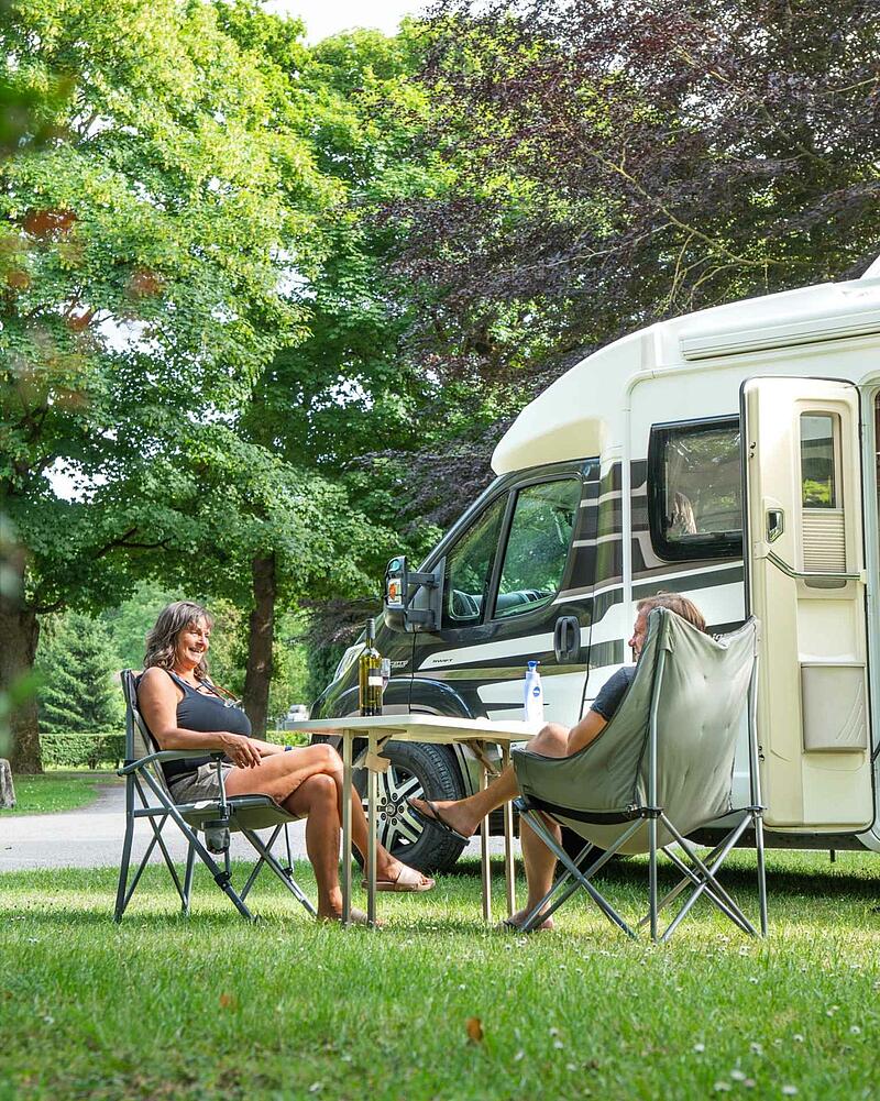 [Translate to Anglais:] A man and a woman sit in comfortable folding chairs at a camping table with a bottle of wine, relaxing next to their modern motorhome on a shaded, grassy pitch.