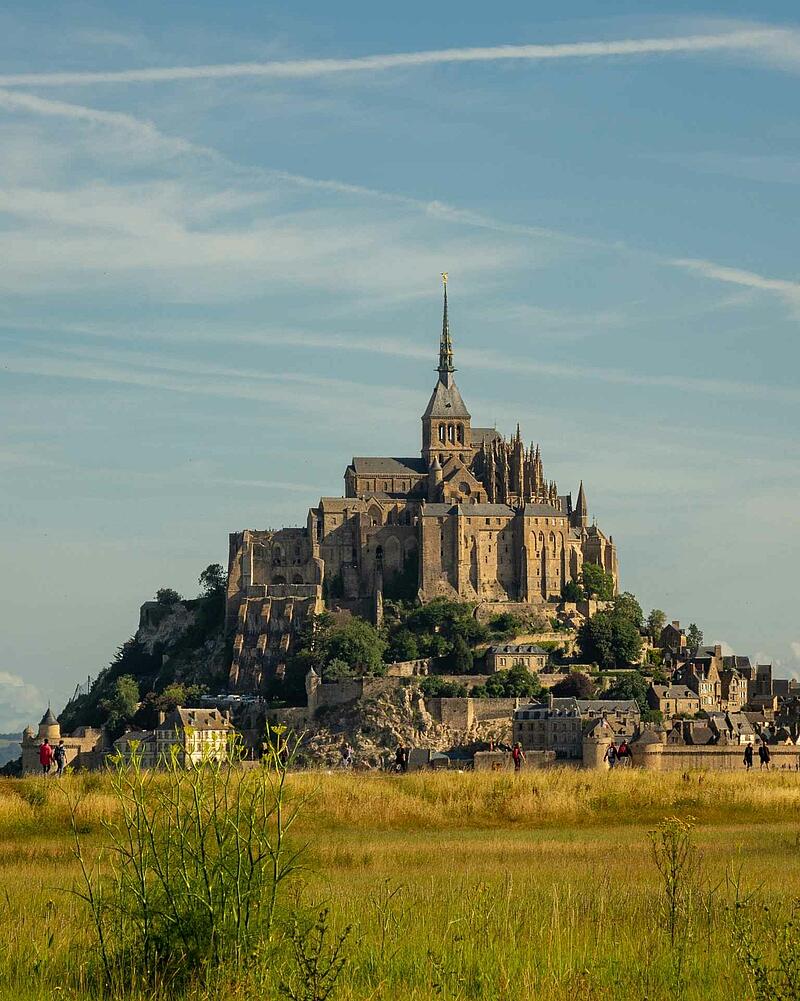[Translate to Anglais:] The magnificent tidal island of Mont-Saint-Michel with its towering medieval abbey, viewed from across a grassy green and yellow field under a light blue summer sky.