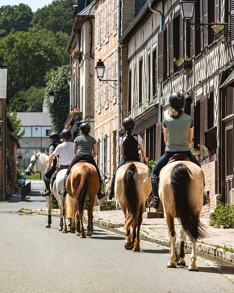 [Translate to Anglais:] A group of riders on horseback walking down a village street lined with classic half-timbered stone houses, an outdoor activity proposed near your Les Castels camping resort.