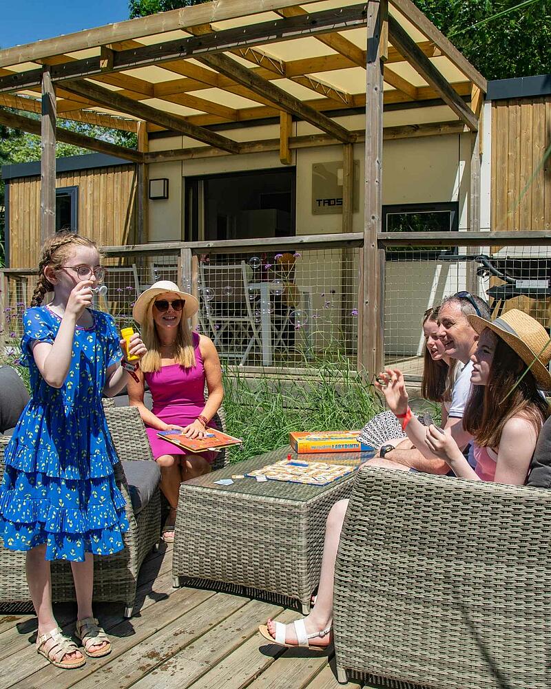 [Translate to Anglais:] A family enjoys quality time together playing a board game and blowing bubbles on the spacious wooden terrace of a premium, modern mobile home at a Les Castels campsite.