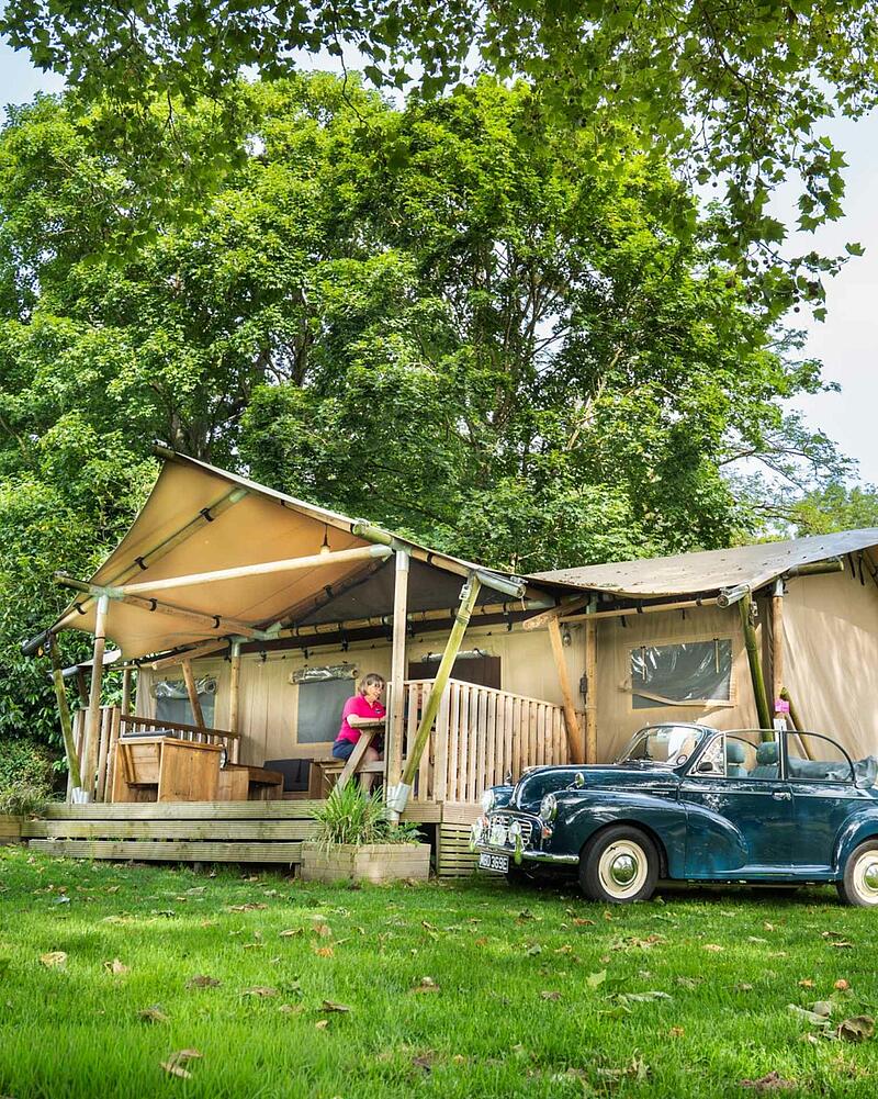 [Translate to Anglais:] A large canvas glamping safari tent with a wooden porch set amongst lush green trees on a premium pitch at a Les Castels 5-star campsite.