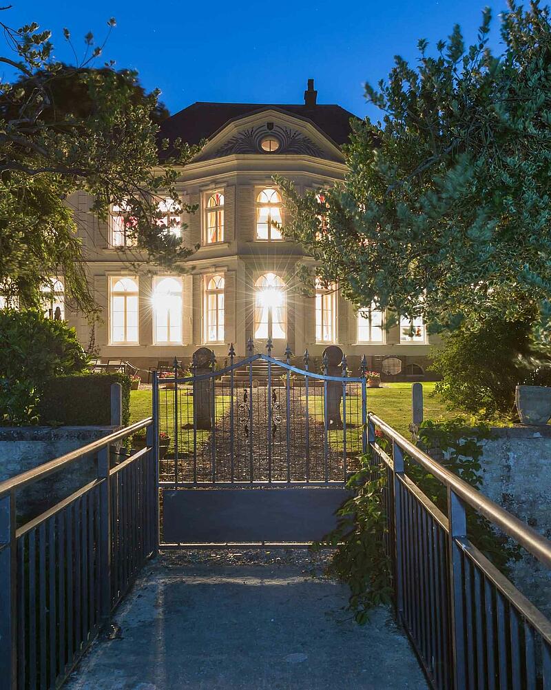 [Translate to Anglais:] The main manor house of Camping Les Castels La Bien-Assise beautifully illuminated at night, viewed from across a small bridge and iron gate under a clear evening sky.