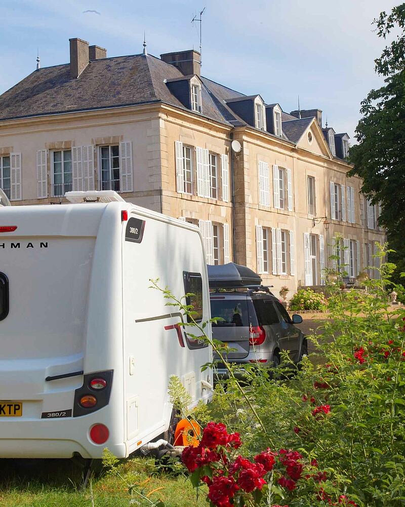 [Translate to Anglais:] The back of a white caravan and a grey SUV parked on a sunny grass pitch in front of a grand, historic stone chateau, beautifully framed by blooming red roses in the foreground.