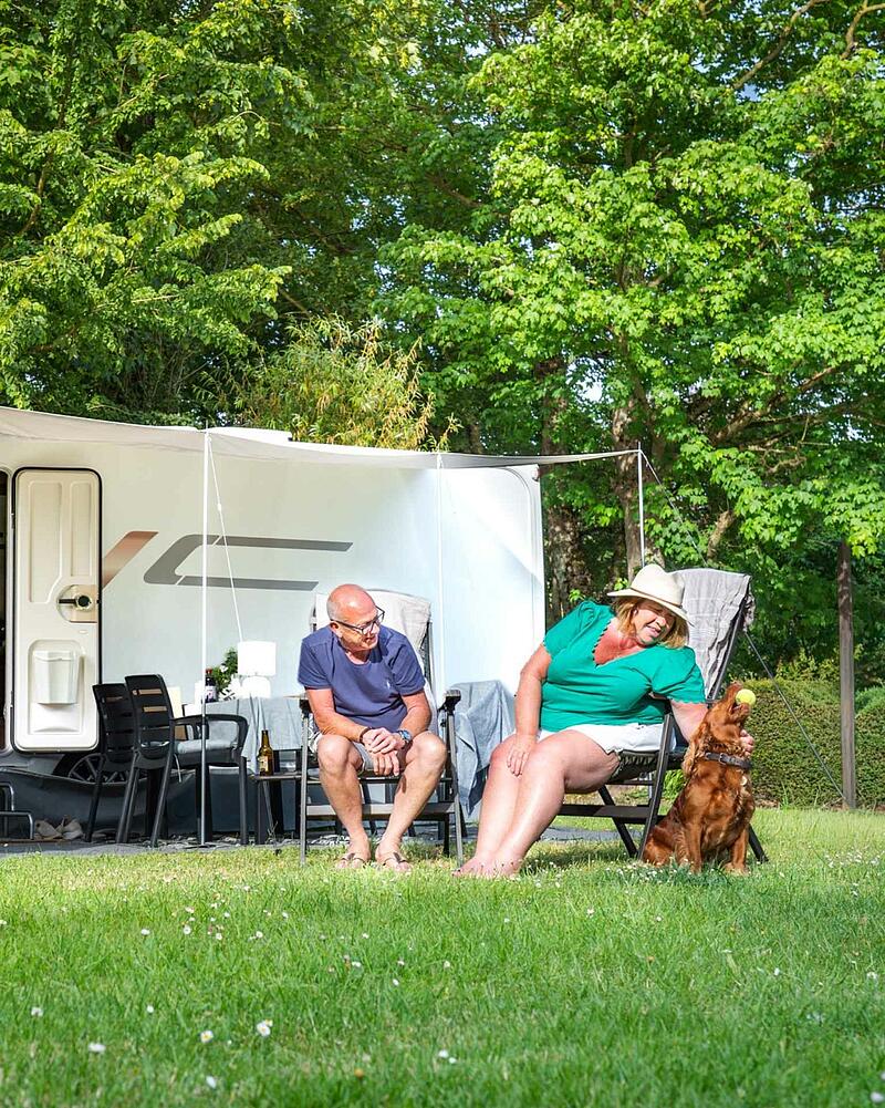 [Translate to Anglais:] A man and woman relaxing in chairs outside their caravan on a lush green lawn, playing with their brown dog holding a tennis ball at a pet-friendly campsite.