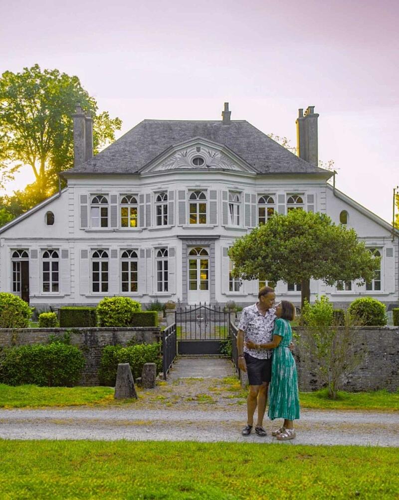 [Translate to Anglais:] A couple standing lovingly in front of a historic French chateau estate at Camping La Bien-Assise during a warm, golden sunset.