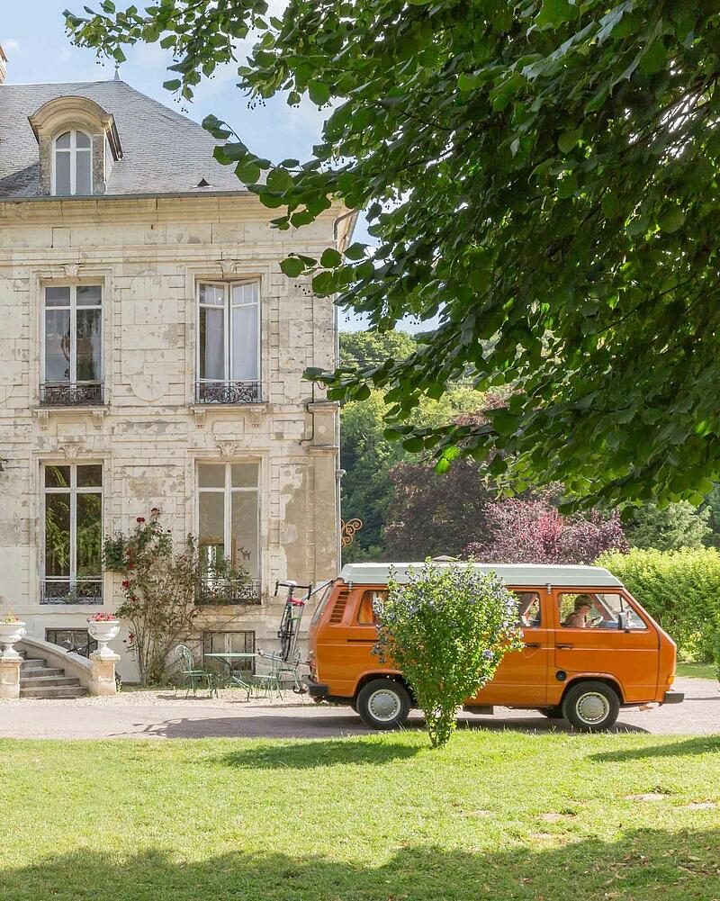 [Translate to Anglais:] A classic orange campervan parked on the grass beside a majestic, historic stone chateau building, perfect for a road trip stopover from Dover to Normandy.