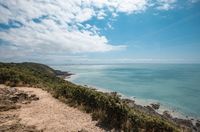 [Translate to Anglais:] A scenic coastal view in Normandy featuring a dirt pathway on a grassy cliff overlooking a vast expanse of turquoise sea under a blue sky with fluffy white clouds, perfect for an outdoor stay at Les Castels.